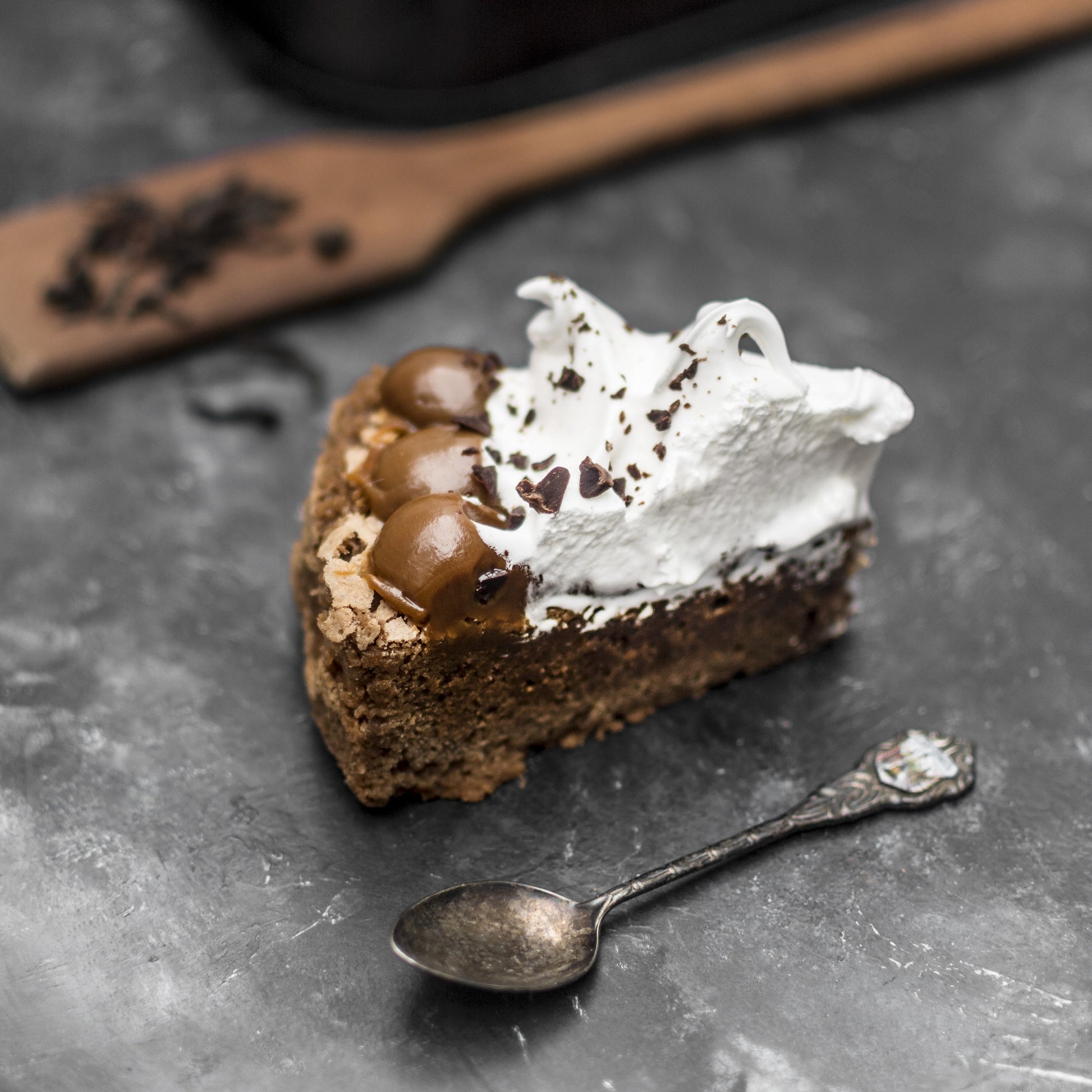 Angled top view of a brownie cake slice crowned with Italian meringue and salted caramel dollops, with a teaspoon, wooden spoon, and other cake slices in the background.