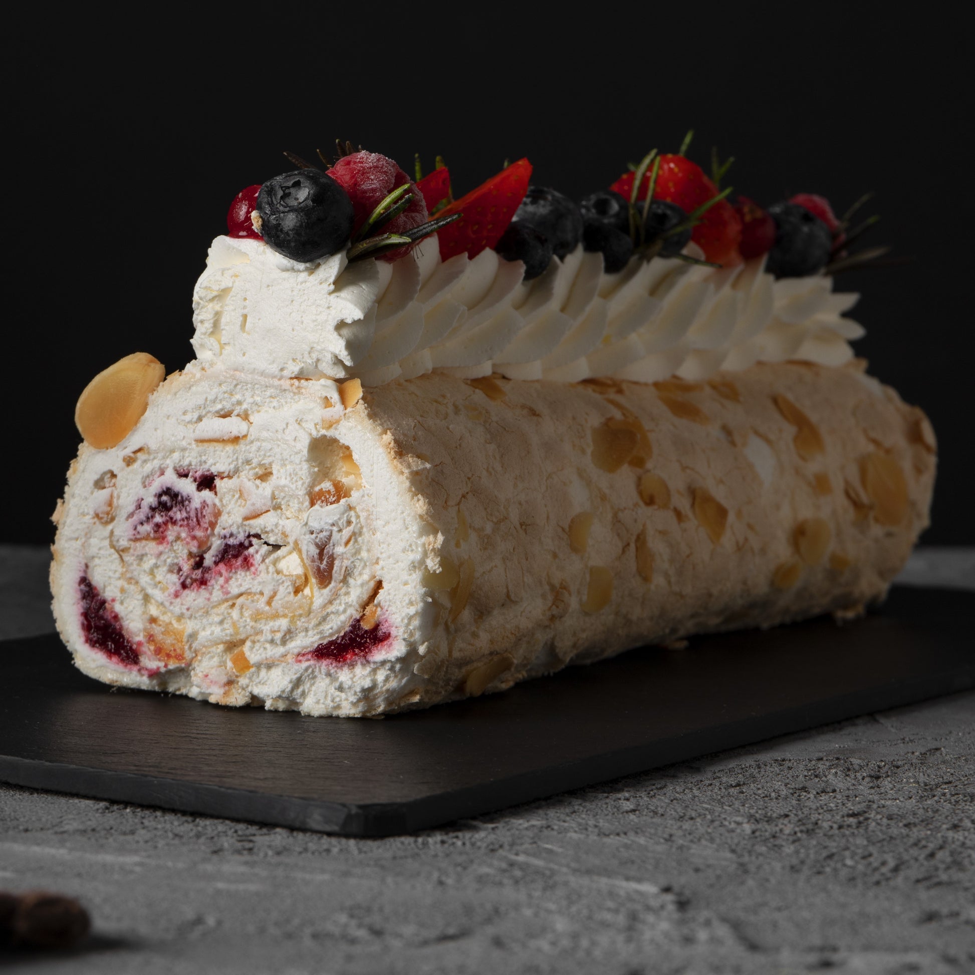Front view of a tropical rolled cake with whipped cream swirls, açai compote filling, fresh berries décor and rosemary leaves, on a black slate board.
