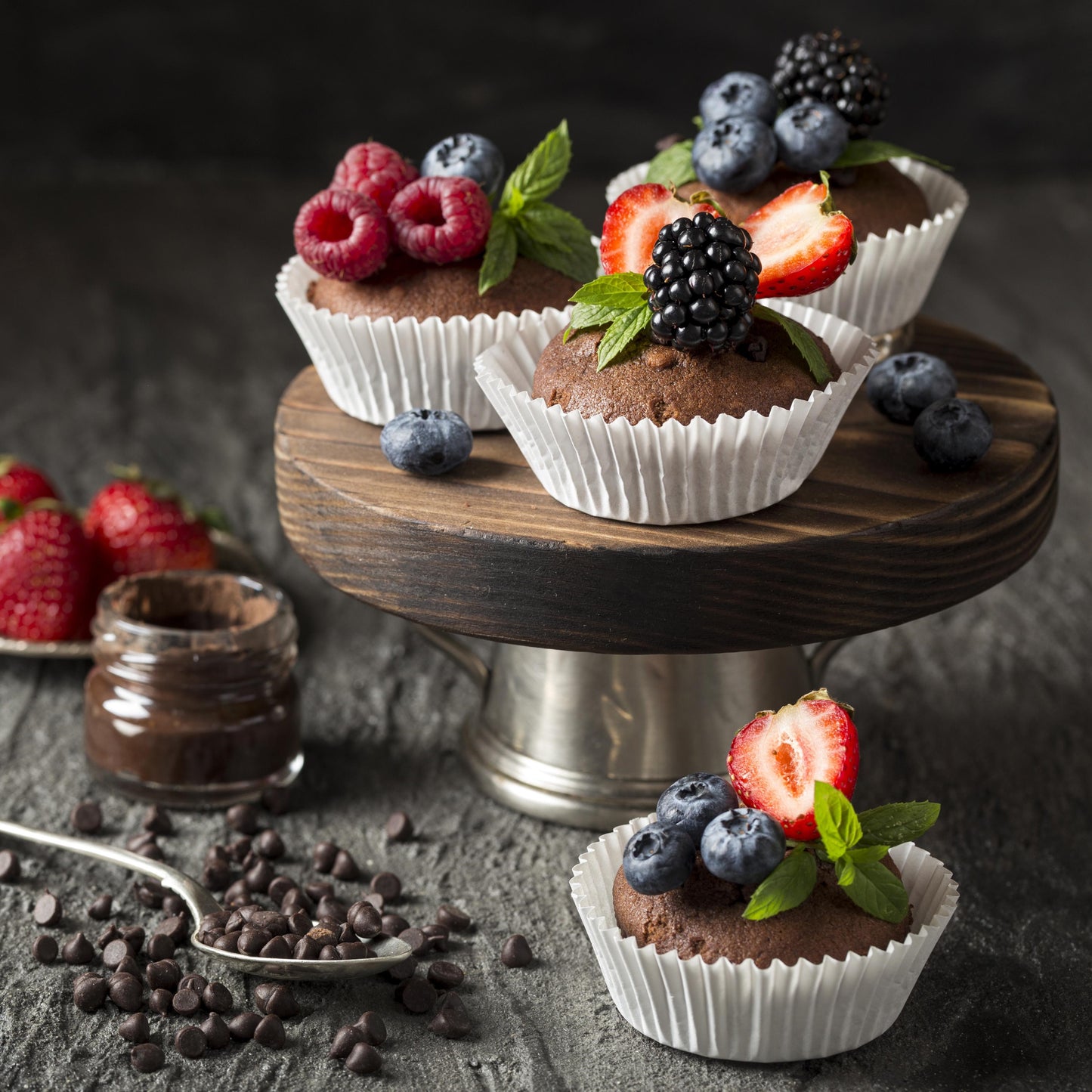 Angled front view of three chocolate chip cupcakes topped with fresh forest fruits and mint leaves, on an elevated round wooden board, with three strawberries, a jar of chocolate spread, a teaspoon, scattered chocolate chips, and another cupcake used as décor in the background.