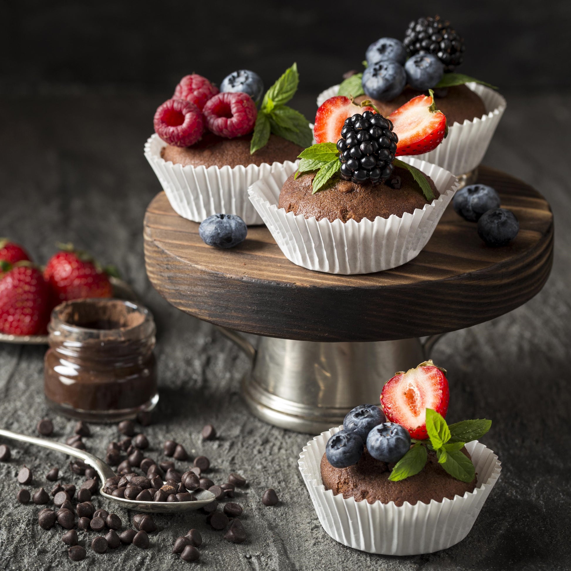 Angled front view of three chocolate chip cupcakes topped with fresh forest fruits and mint leaves, on an elevated round wooden board, with three strawberries, a jar of chocolate spread, a teaspoon, scattered chocolate chips, and another cupcake used as décor in the background.