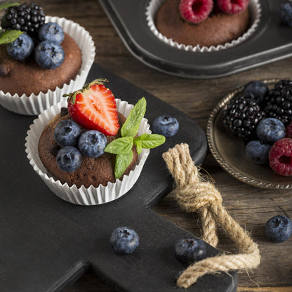 Angled top view of several chocolate chip cupcakes topped with fresh forest fruits and mint leaves, on a black wooden board, with fresh forest fruits, and a tray of muffins used as décor in the background.