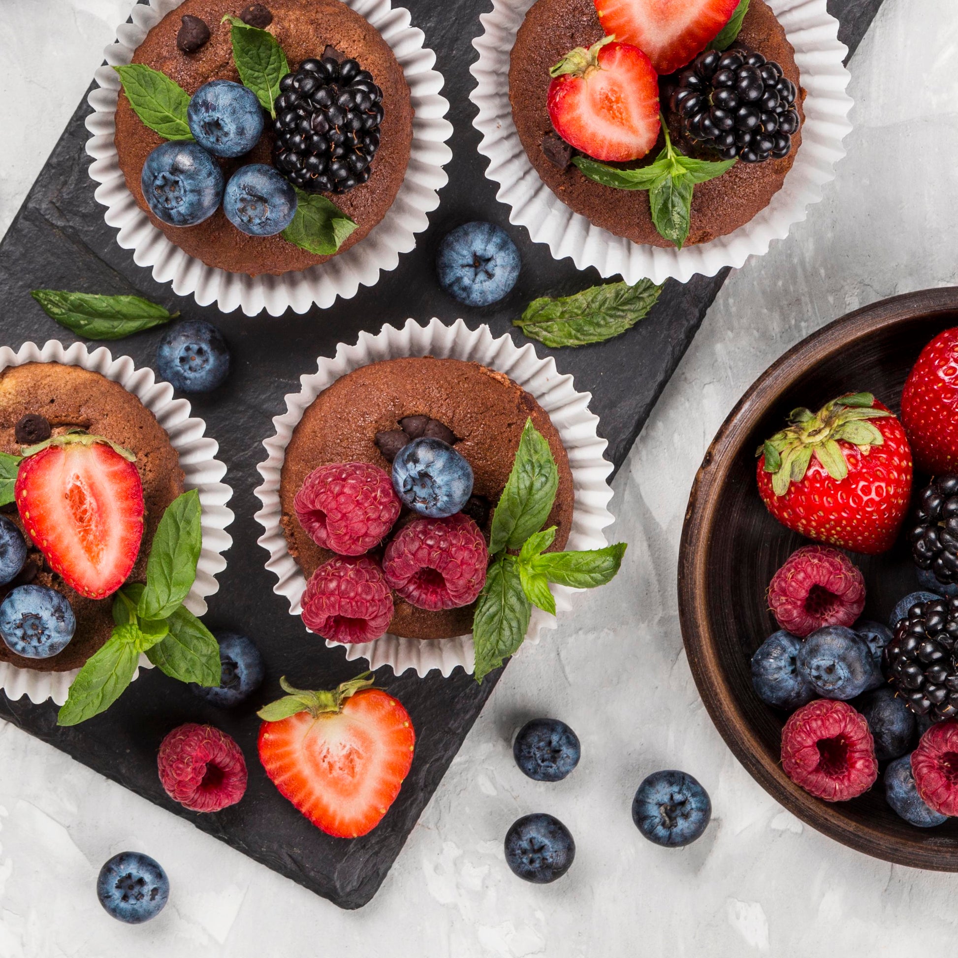 Top view of top view of several chocolate chip cupcakes topped with fresh forest fruits and mint leaves, on a black slate board, with a bowl of fresh forest fruits and scattered berries used as décor in the background.