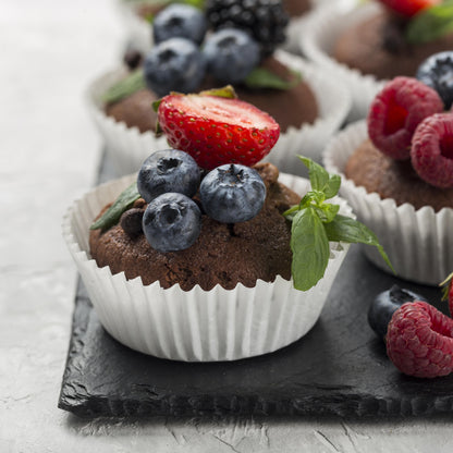 Front close-up view of several chocolate chip cupcakes topped with fresh forest fruits and mint leaves, on a black slate board.