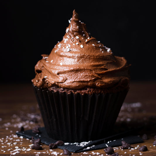 Front close-up view of a chocolate giant cupcake decorated with a mix of hazelnut-chocolate buttercream and topped with coconut flakes, on a black cloth with scattered chocolate chips, and coconut flakes used as décor in the background.