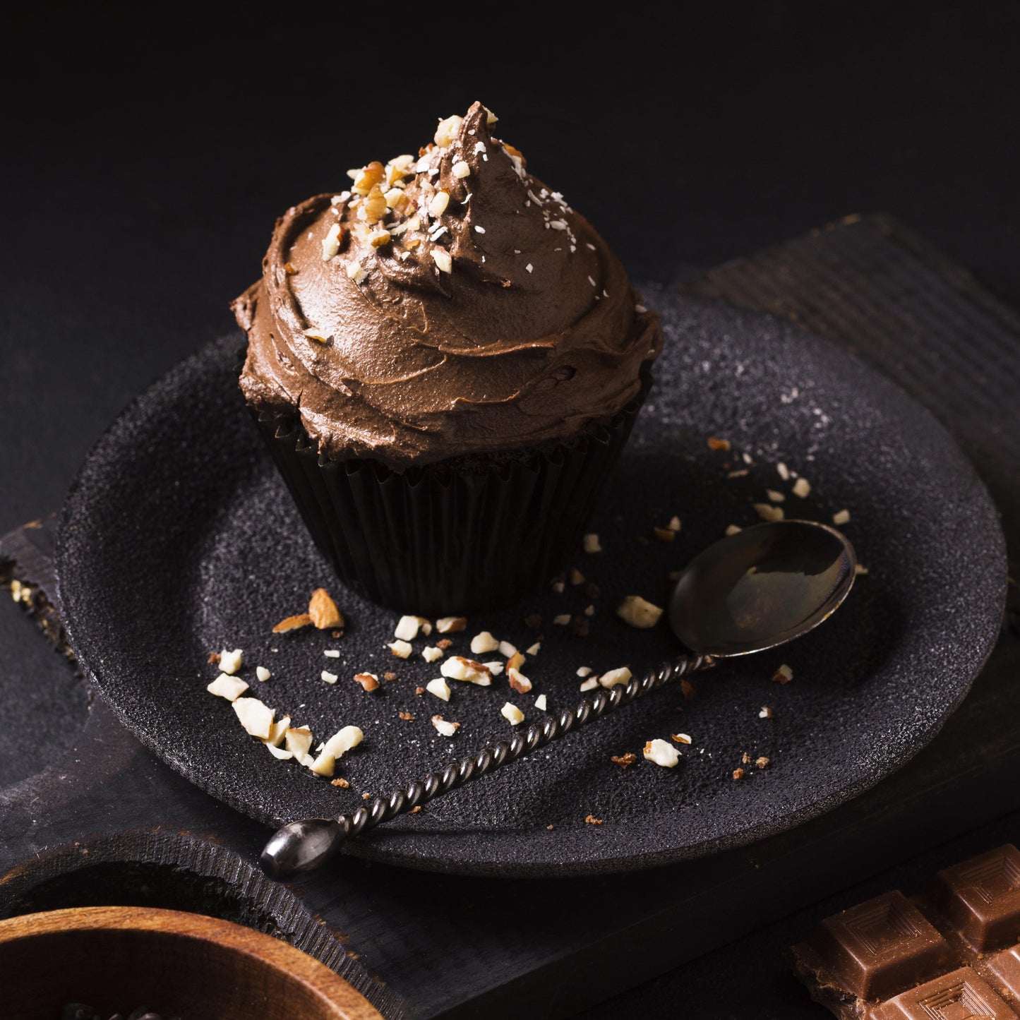 Angled top view of a chocolate giant cupcake decorated with a mix of hazelnut-chocolate buttercream, topped with roasted hazelnut crisps and coconut flakes, on a black plate, with scattered hazelnuts, a teaspoon, a wooden bowl of chocolate chips, and broken chocolate chunks used as décor in the background. 