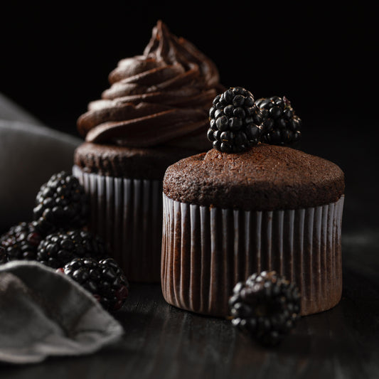 Angled front close-up view of two tall chocolate ramekin-shaped cupcakes, one decorated with chocolate buttercream and the other topped with fresh blackberries.