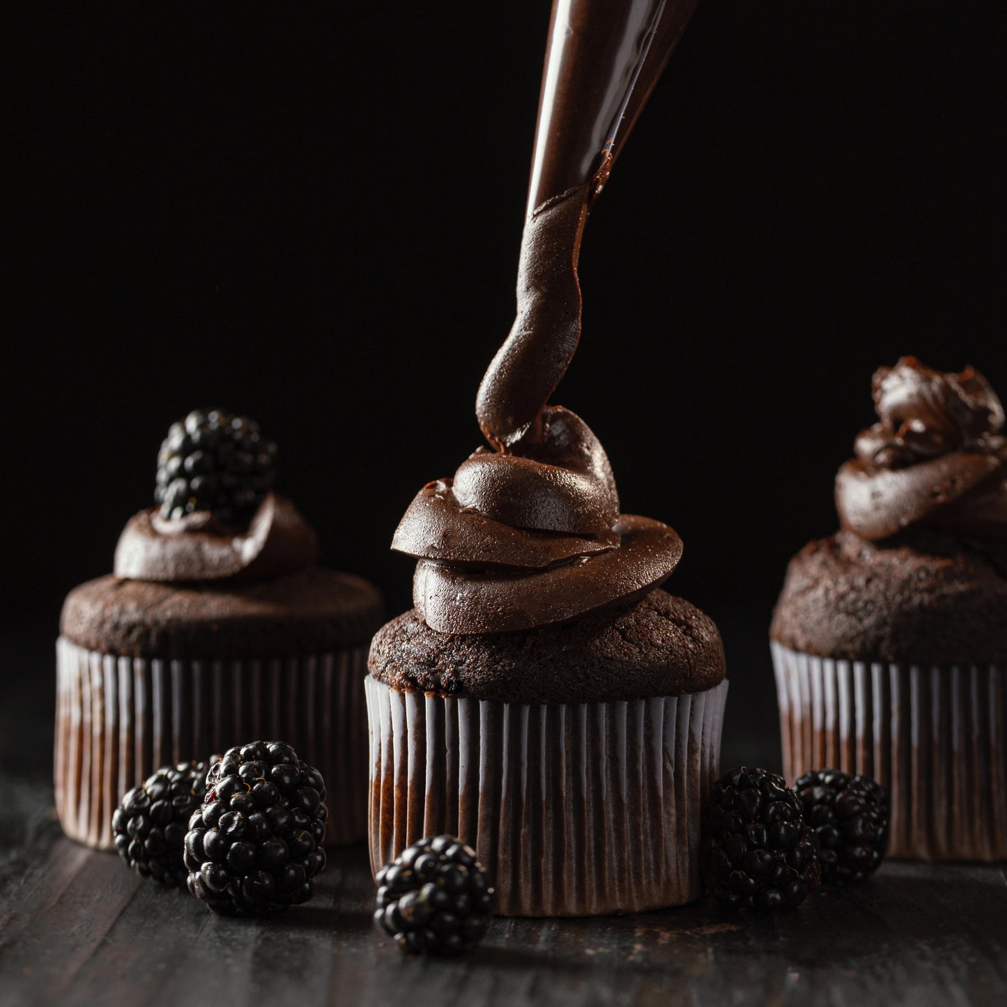 Front view of of three tall chocolate ramekin-shaped cupcakes, one being piped with chocolate buttercream, one already decorated with chocolate buttercream, and one topped with fresh blackberries.