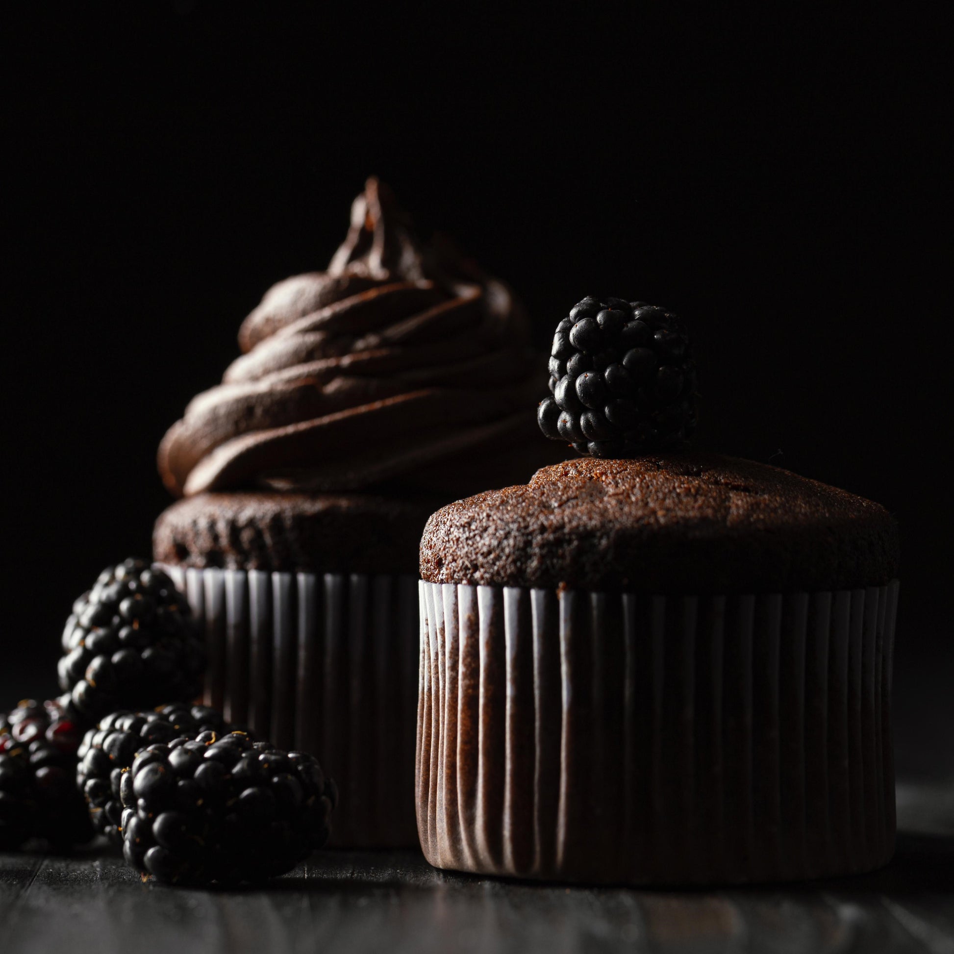 Front close-up view of two tall chocolate ramekin-shaped cupcakes, one decorated with chocolate buttercream and the other topped with fresh blackberries.