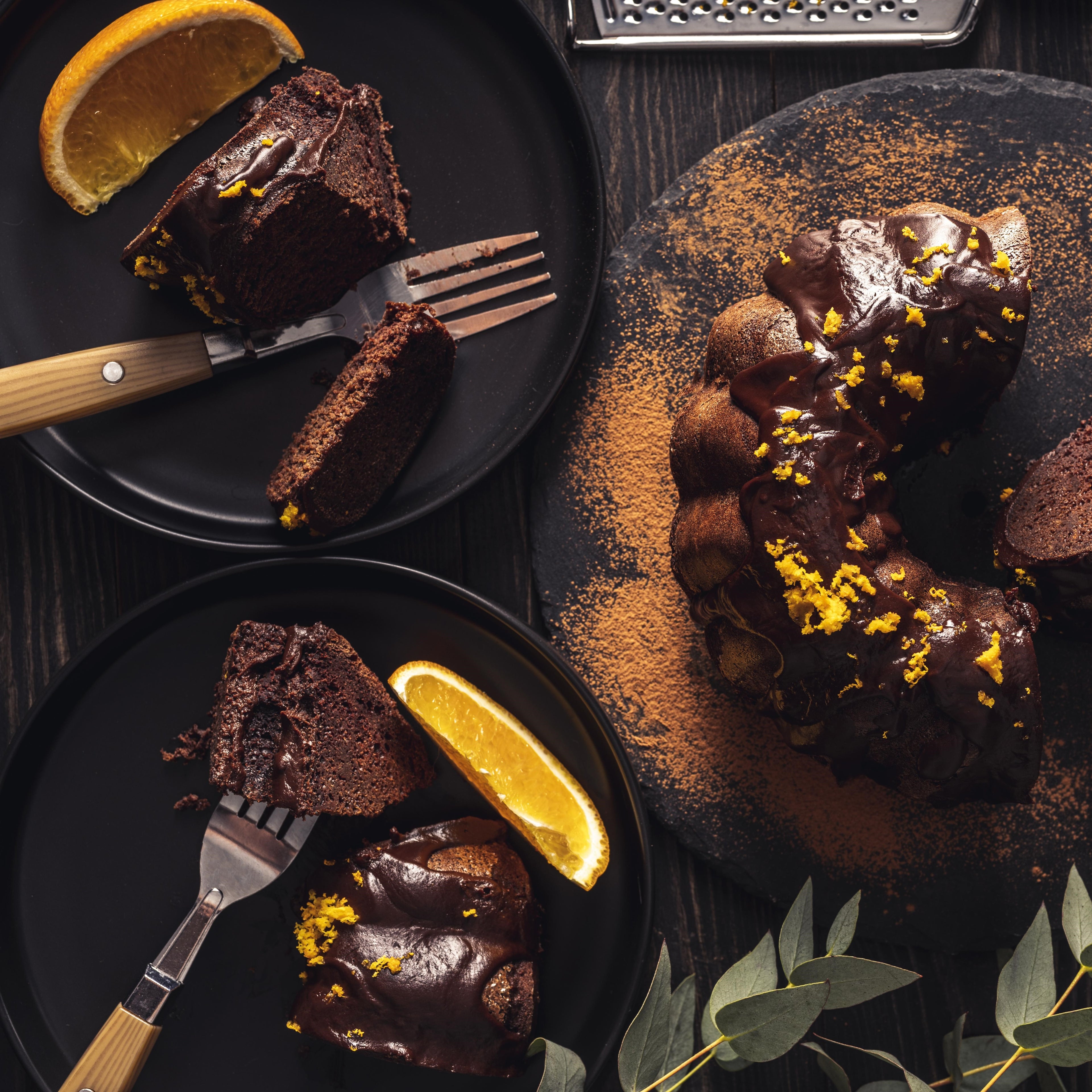 Top view of a cut chocolate Bundt cake drizzled with chocolate ganache and orange zest next to two black plates with slices of Bundt cake, forks, and orange wedges.